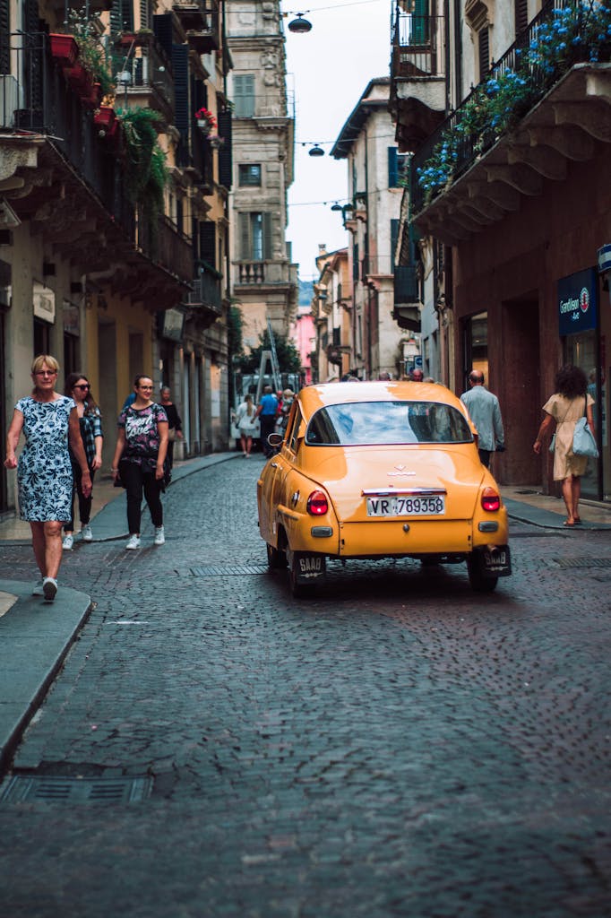 Charming scene of a vintage yellow car driving down a cobblestone street in Ferrazze, Italy, with pedestrians walking nearby.
