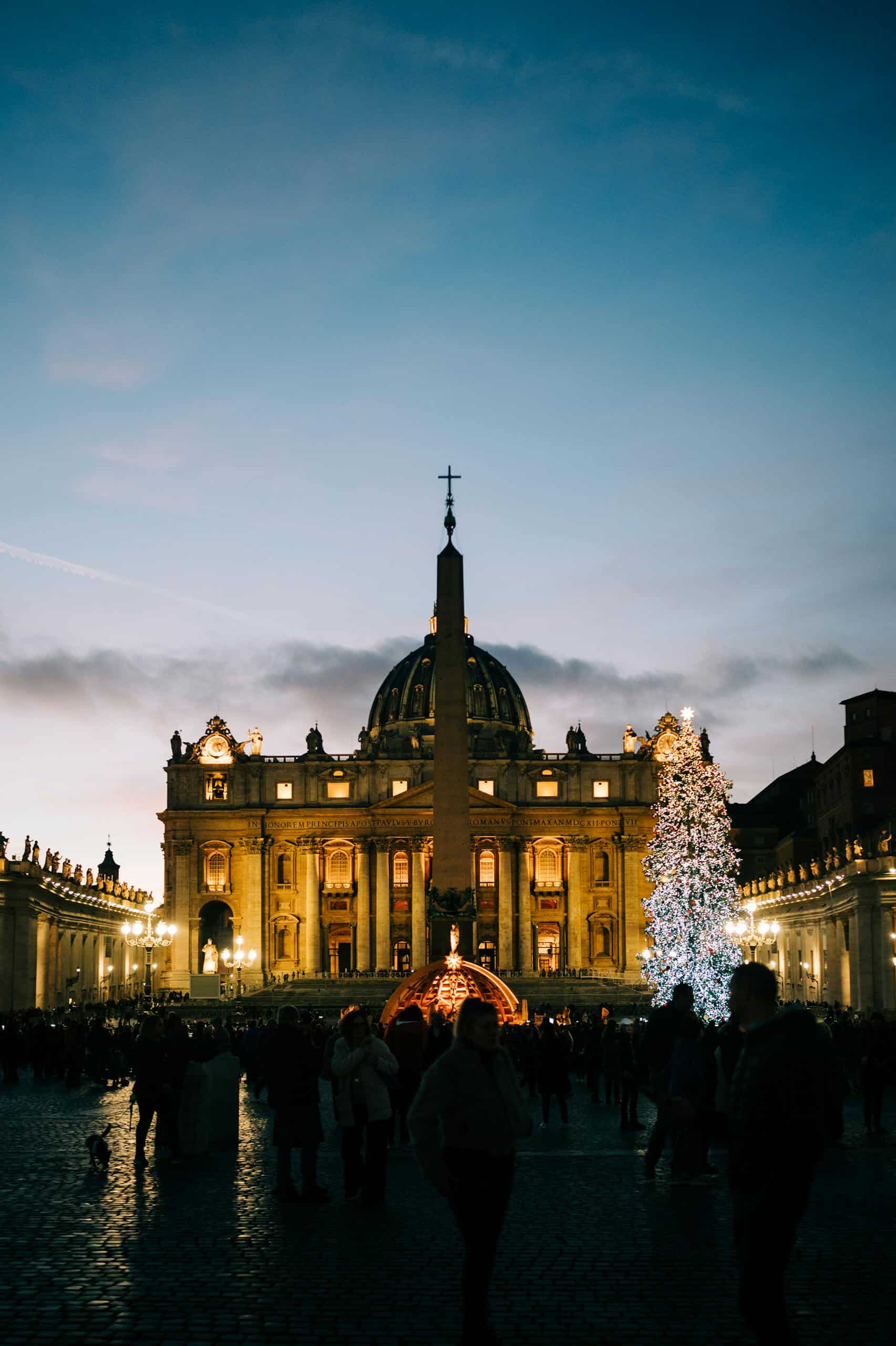 Illuminated St. Peter's Basilica with Christmas tree and crowd at night in Vatican City.