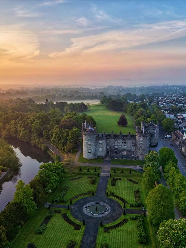 Stunning aerial view of medieval Kilkenny Castle, Ireland at sunrise. Lush gardens and river visible.