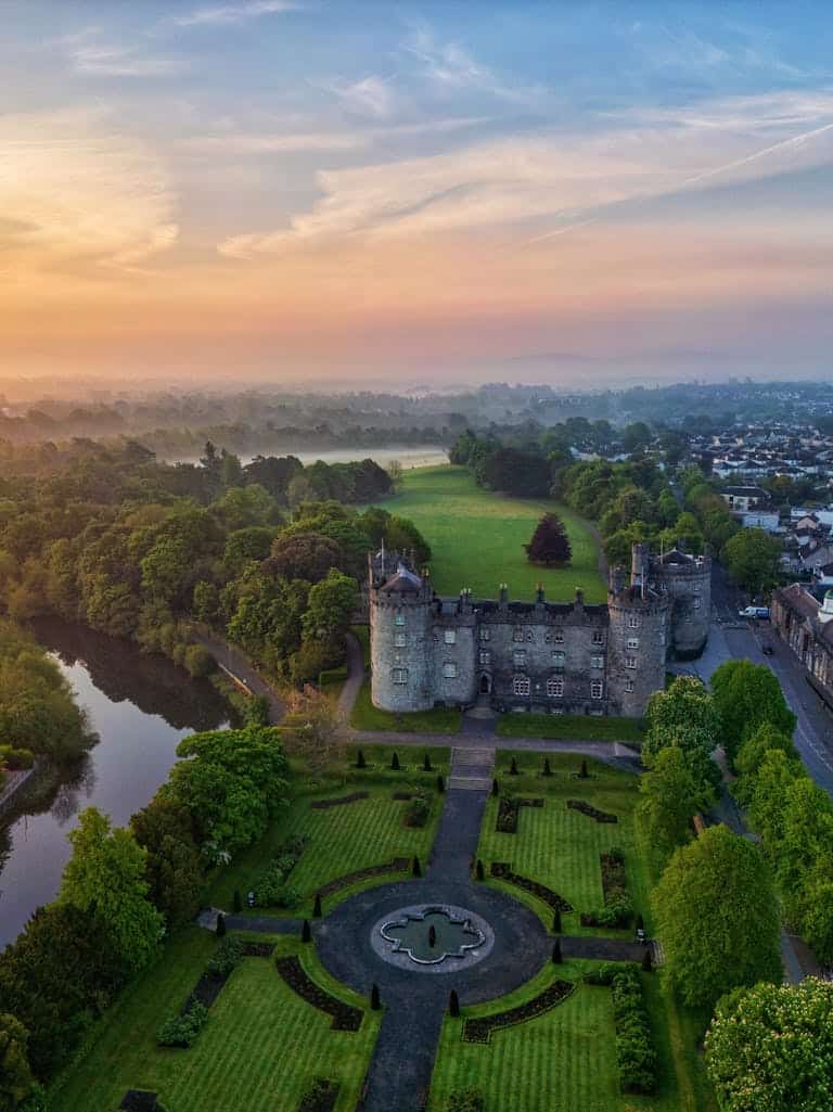 Stunning aerial view of medieval Kilkenny Castle, Ireland at sunrise. Lush gardens and river visible.