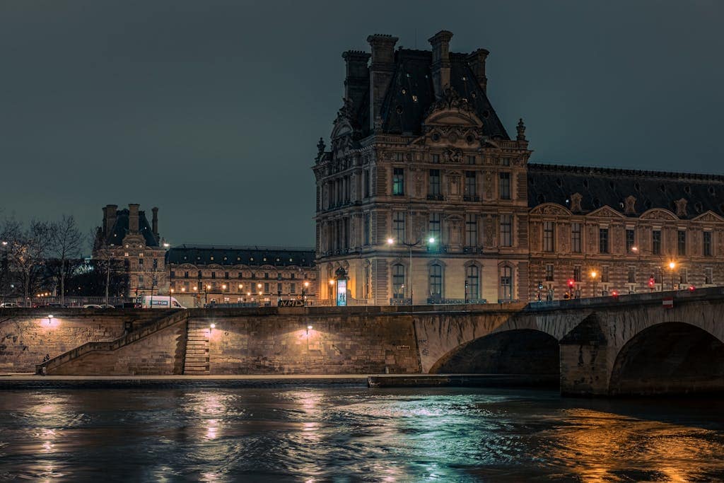 Illuminated Louvre Museum by the Seine in Paris at night, showcasing historic architecture.