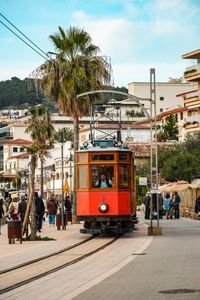 Classic red tram travels through the bustling streets of Soller, Mallorca, Spain, amid tourists and vibrant palm trees.